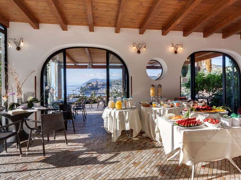 Ein Frühstücksbuffet mit Obst, Müsli und Säften wird in einem Speisesaal mit großen Fenstern und Blick auf das Meer und eine Stadt angeboten.