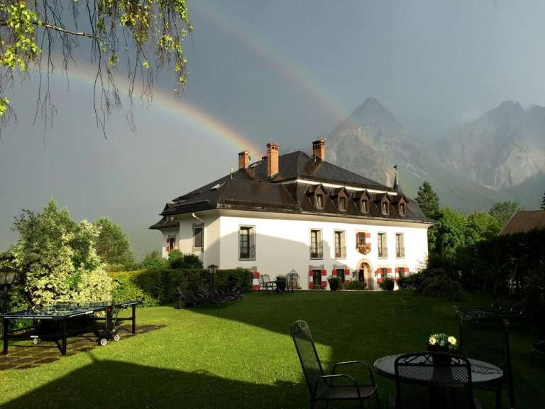 Ein großes, weißes Haus mit roten Fensterläden steht in einem Garten mit Bergen im Hintergrund und einem Regenbogen am Himmel.