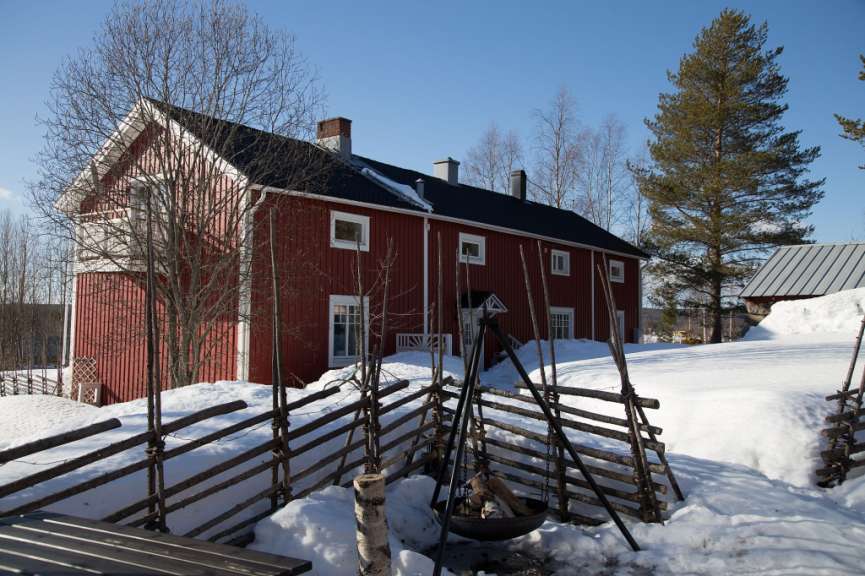 Eine rote Holzhütte mit Schnee bedecktem Dach und Zaun davor steht an einem sonnigen Tag im Winter.