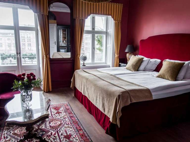 Ein luxuriös eingerichtetes Hotelzimmer mit einem großen Bett, roten Wänden und einem Fenster mit Blick auf die Stadt.