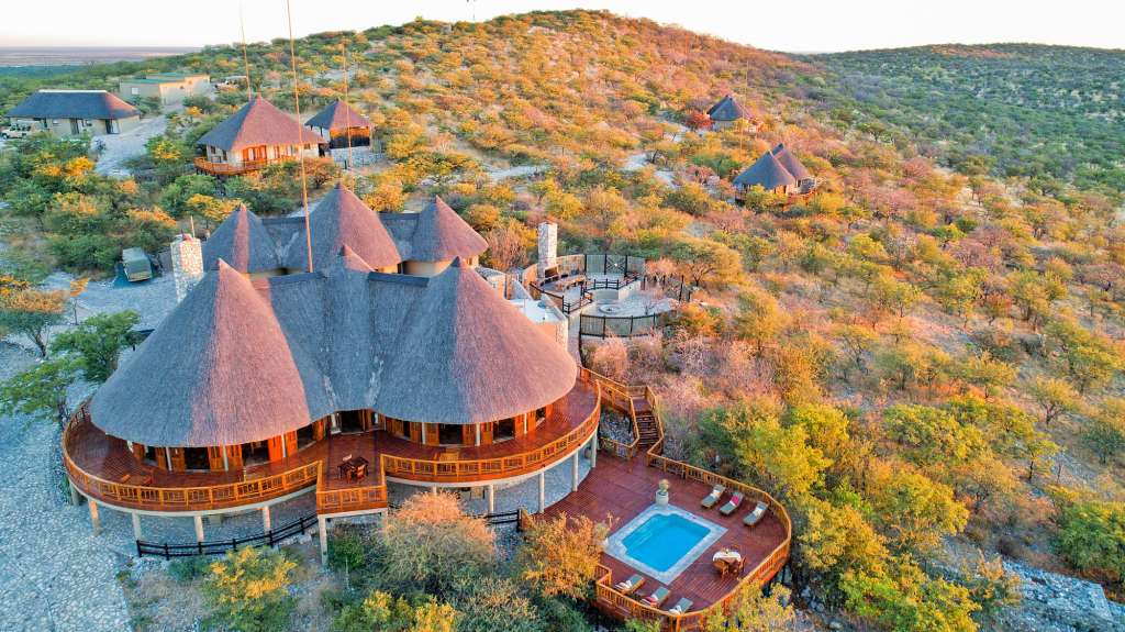 Eine malerische Drohnenaufnahme zeigt ein Safari-Camp mit mehreren traditionellen Strohdach-Bungalows, die auf einem hügeligen Gelände in der goldenen Stunde der Abendsonne liegen.
