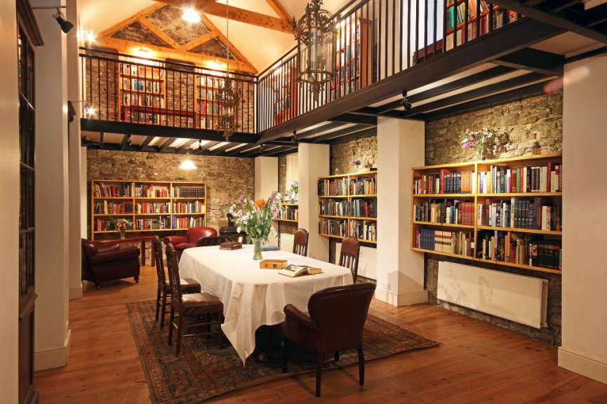 Eine Innenaufnahme zeigt ein gemütliches Leseraum mit Bücherregalen, einem Esstisch mit weißer Tischdecke, mehreren Stühlen und zwei roten Sesseln.