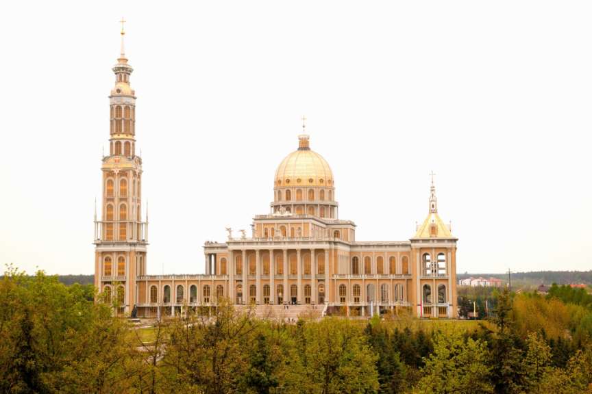 Dieses Bild zeigt die imposante Basilika Unserer Lieben Frau von Licheń in Polen, mit ihrem hohen Turm, der goldenen Kuppel und dem weitläufigen Gebäude, eingerahmt von Bäumen.