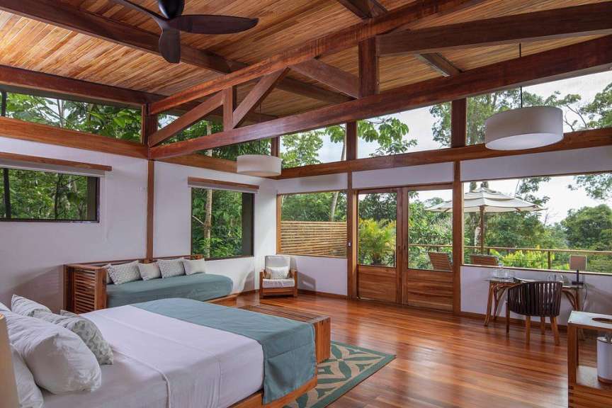 Ein Schlafzimmer mit Holzbalkendecke und großen Fenstern, die einen Blick auf den Regenwald freigeben.