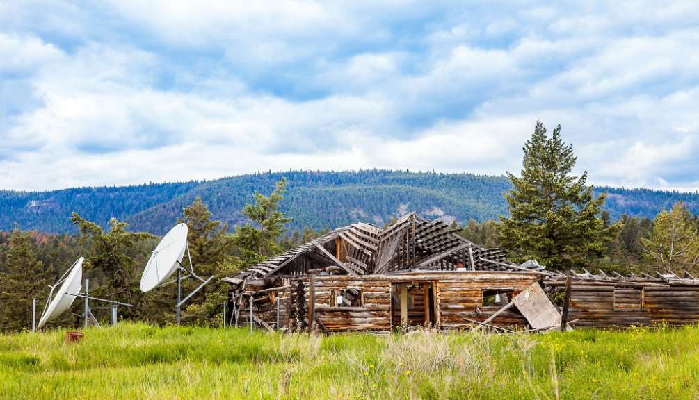Ein verfallenes Blockhaus steht in einer grünen Wiese unter einem bewölkten Himmel vor einem von Bäumen bedeckten Hügel.