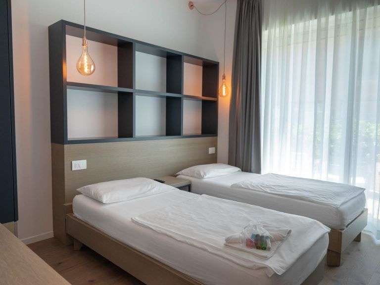 Ein modernes Hotelzimmer mit zwei Einzelbetten und einem Regal über den Betten wird durch ein Fenster mit Vorhängen beleuchtet.