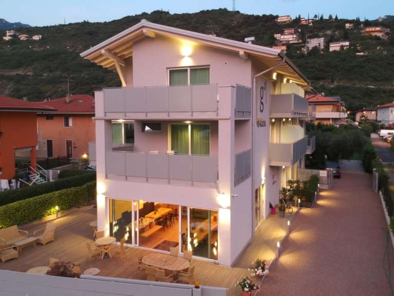 Ein modernes, mehrstöckiges Gebäude mit Balkonen und einer Holzterrasse steht abends in einer hügeligen Landschaft.