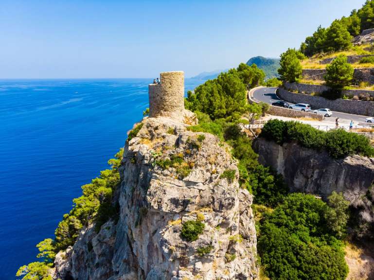 Ein verwitterter Turm aus Stein steht auf einem felsigen Vorsprung über dem türkisfarbenen Meer, während eine kurvenreiche Straße mit Autos an einem Hang mit üppiger Vegetation entlangführt.