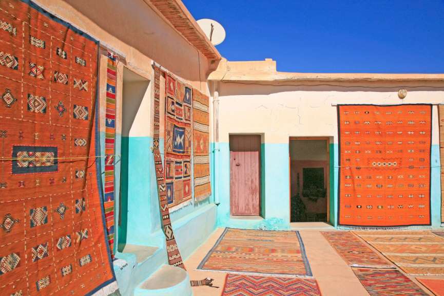 Mehrere bunte Teppiche sind an einer hellblauen und beigen Wand auf einem sonnigen Innenhof ausgestellt.