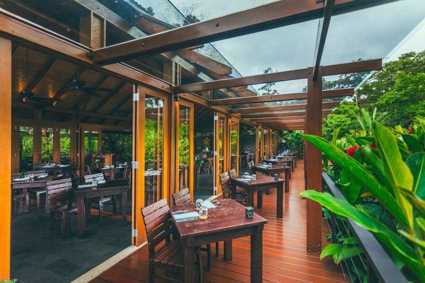 Ein Außenrestaurant mit Holztischen und Stühlen unter einem Glasdach, umgeben von üppiger Vegetation.