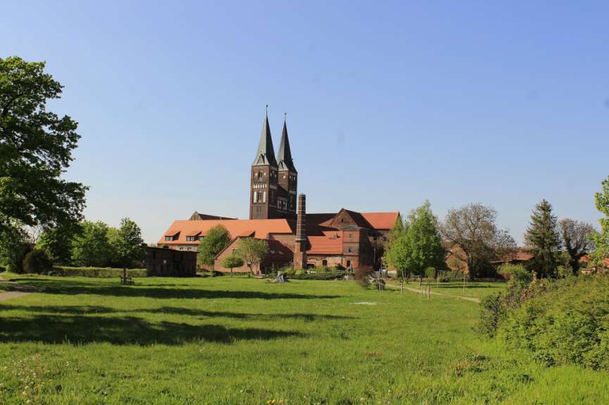 Ein Backsteingebäude mit zwei hohen Türmen steht auf einer grünen Wiese unter klarem Himmel.