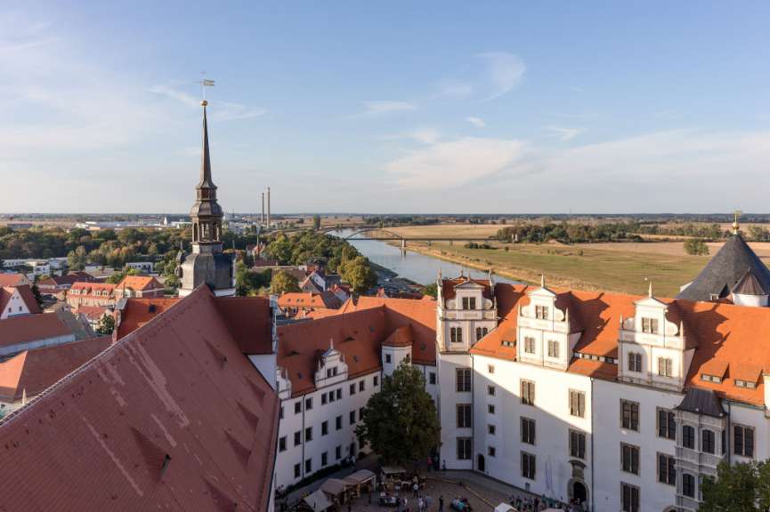 Ein Luftbild zeigt eine Stadtansicht mit einem Kirchturm und verschiedenen Gebäuden, die sich bis zu einem Fluss und darüber hinaus erstrecken.