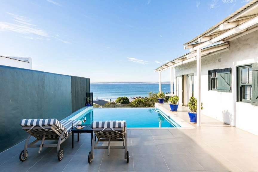 Ein Infinity-Pool und zwei Liegestühle stehen auf einer gepflasterten Terrasse mit Blick auf das Meer und ein weißes Haus mit grünen Fensterläden.