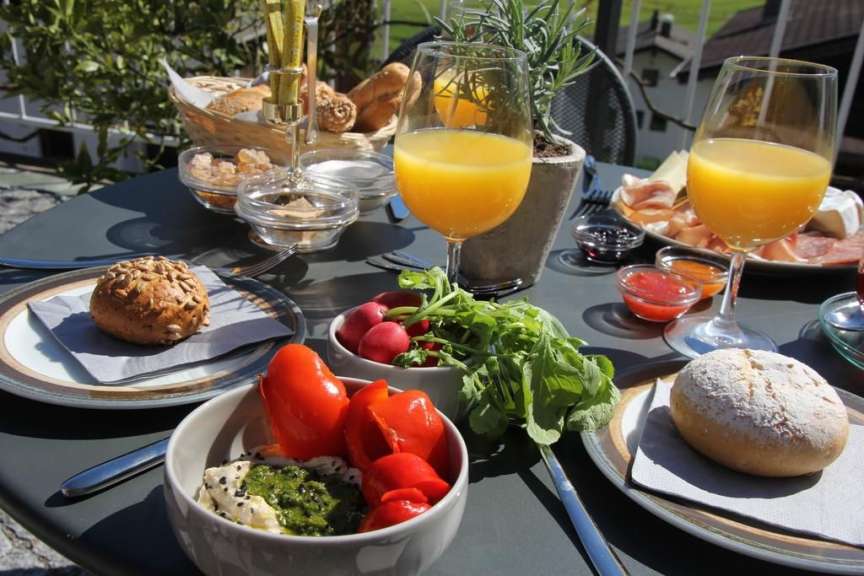 Auf einem Tisch im Freien steht ein Frühstück mit Orangensaft, Gebäck, Aufschnitt, Käse, Radieschen und Dip.