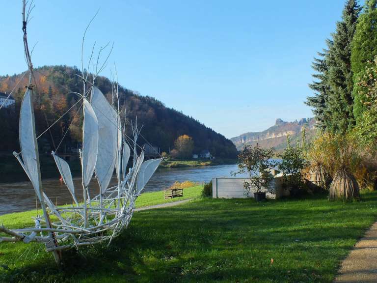 Eine weiße, abstrakte Skulptur in Form eines Schiffes steht am Ufer eines Flusses mit Blick auf eine bewaldete Hügellandschaft unter einem klaren blauen Himmel.