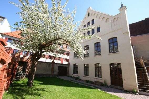 Ein blühender Baum steht vor einem Gebäude mit mehreren Fenstern und einer Treppe, das an einem sonnigen Tag von der Seite aufgenommen wurde.