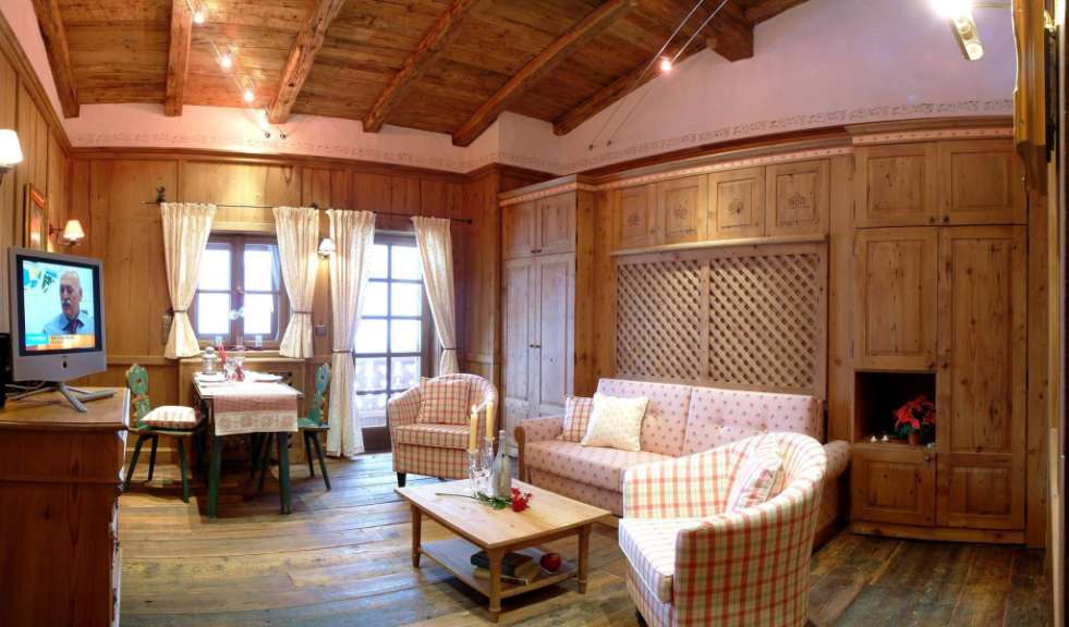 In einem gemütlichen, holzgetäfelten Zimmer mit Holzbalkendecke und rustikaler Einrichtung stehen ein Sofa, zwei Sessel, ein Couchtisch, ein kleiner Esstisch mit Stühlen und ein Fernseher.