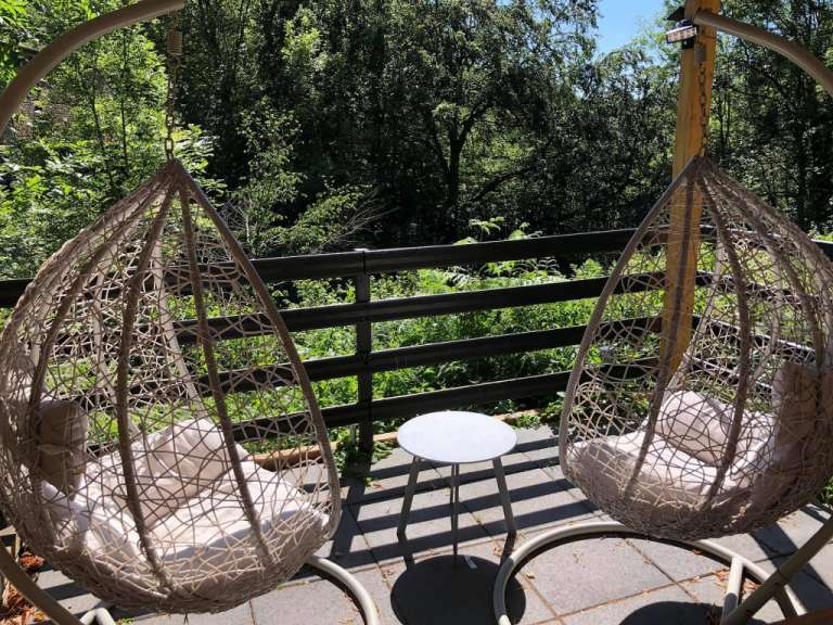 Zwei Hängeschaukeln mit Kissen stehen auf einer Terrasse vor einer grünen Hecke.