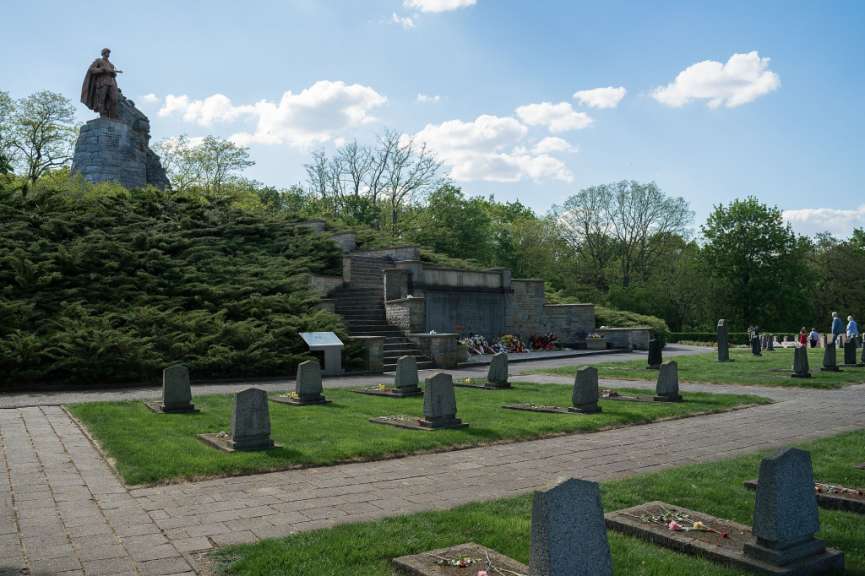 Eine Gruppe von Menschen geht über einen Friedhof, der von einem großen Denkmal mit einer Statue dominiert wird.