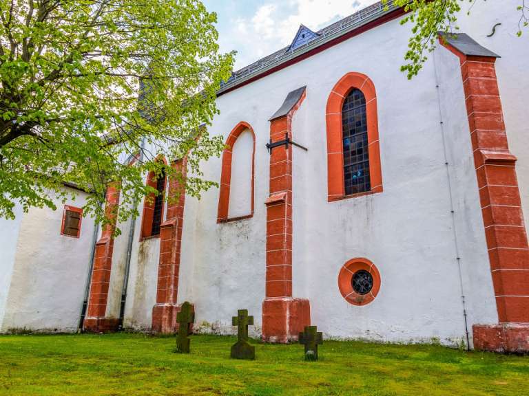 Eine Kirche mit weißen Wänden und roten Zierelementen steht auf einer grünen Rasenfläche, auf der mehrere Grabkreuze zu sehen sind.