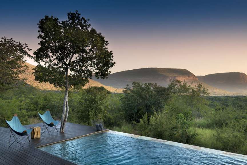 Ein Infinity-Pool mit Blick auf eine hügelige Landschaft bei Sonnenuntergang, flankiert von zwei Liegestühlen auf einer Holzterrasse.