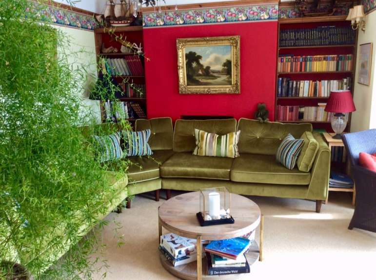 Ein grünes Samtsofa mit bunten Kissen steht vor einem roten Bücherregal in einem Wohnzimmer mit einem Holztisch in der Mitte.