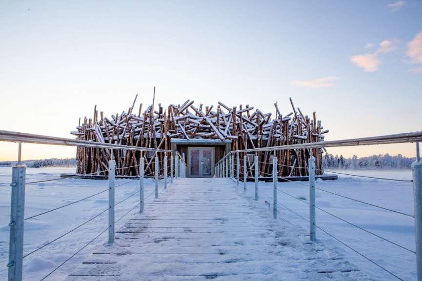 Ein Holzsteg führt zu einem Gebäude aus Holzstäben