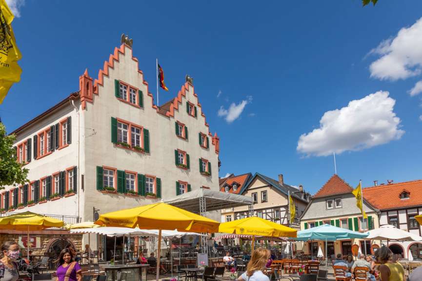 Im Sommer sind Menschen unter Sonnenschirmen in einem Freiluftcafé in einer Stadt mit historischen Gebäuden und blauem Himmel zu sehen.