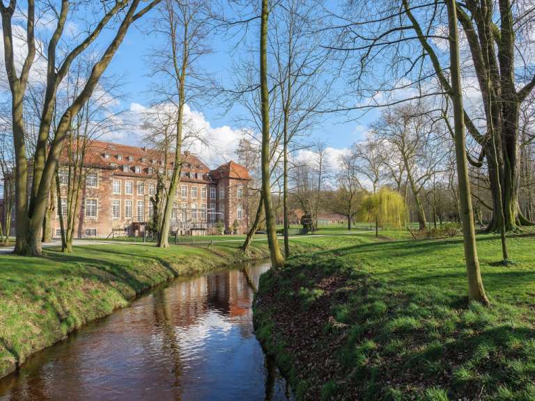 Ein Fluss fließt durch einen Park mit Bäumen und einem Gebäude im Hintergrund.