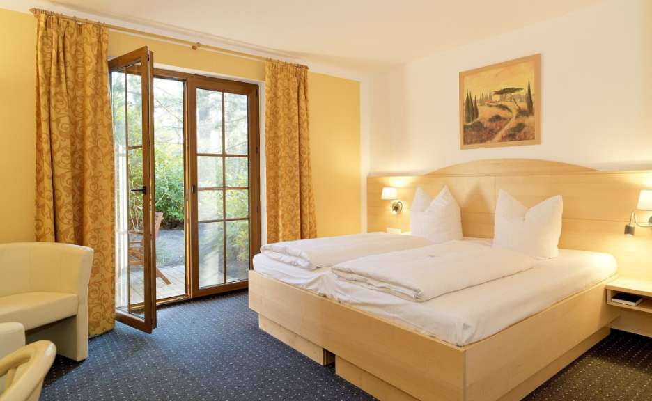 Dieses Bild zeigt ein Doppelzimmer mit zwei Betten, das mit einem Holzbettgestell, einer Nachttischlampe, einem Bild über dem Bett und einem Fenster mit Vorhängen ausgestattet ist.