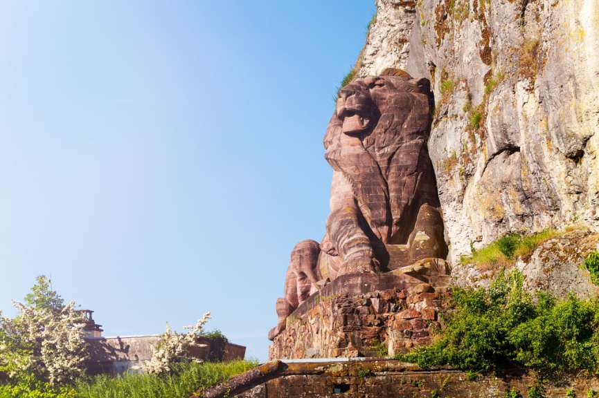 Eine massive, in Fels gehauene Löwenskulptur ragt vor einem blauen Himmel empor, umgeben von grüner Vegetation und Felsen.