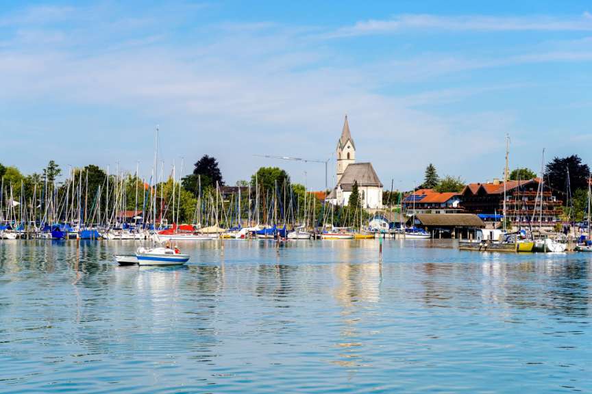 Ein Boot fährt im Vordergrund auf einem ruhigen Gewässer, während im Hintergrund eine Reihe von Segelbooten an einem Steg vertäut ist und eine Stadt mit Kirche und Gebäuden zu sehen ist.