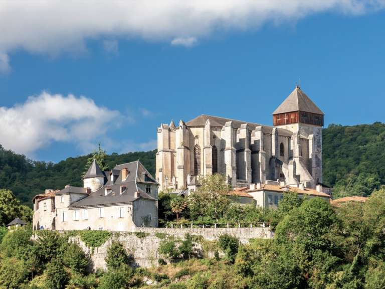Eine historische Kirche mit einem quadratischen Turm und einer Spitze, die von mehreren kleineren Gebäuden und viel Grün umgeben ist, steht unter einem blauen Himmel mit Wolken.