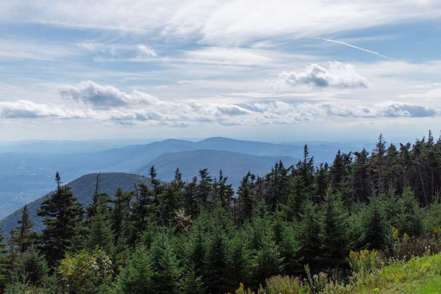 Ein Bergpanorama mit einem Wald im Vordergrund und mehreren blauen Hügelketten im Hintergrund unter einem leicht bewölkten Himmel.