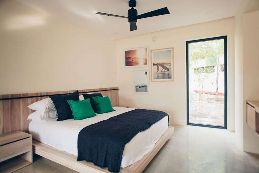 Ein Schlafzimmer mit einem Kingsize-Bett, zwei grünen und zwei schwarzen Kissen, einer dunklen Decke, drei gerahmten Bildern mit Surfmotiven und einem Deckenventilator.