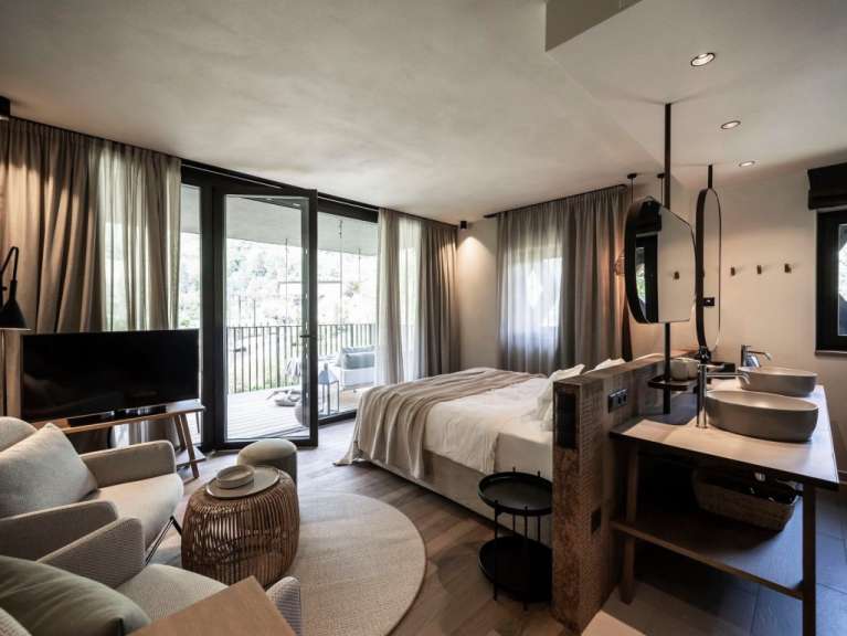 Ein luxuriöses Schlafzimmer mit Doppelbett, Sitzbereich, integriertem Badezimmer und Balkon mit Blick ins Grüne.