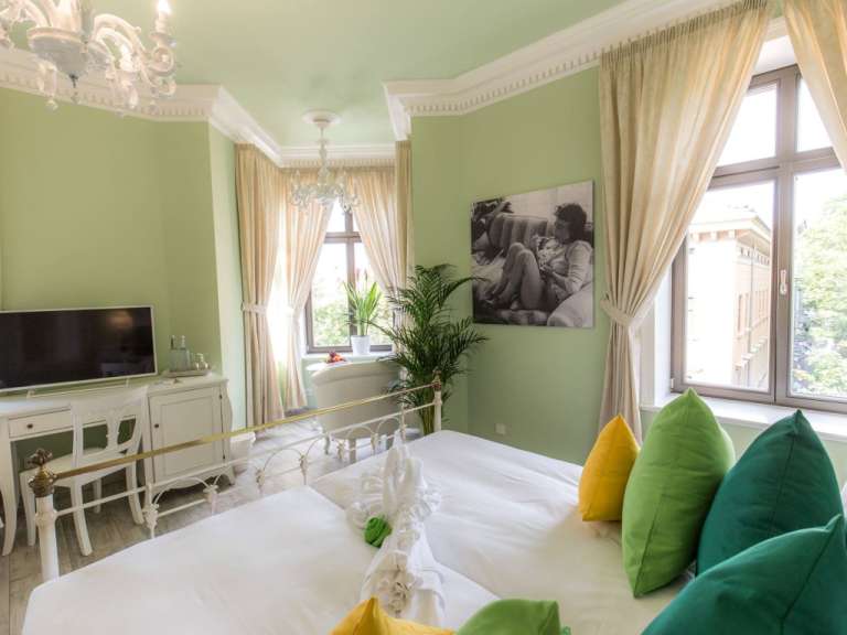 In einem hellgrün gestrichenen Zimmer mit Kronleuchtern, Fernseher und großem Fenster steht ein weißes Bett mit bunten Kissen.