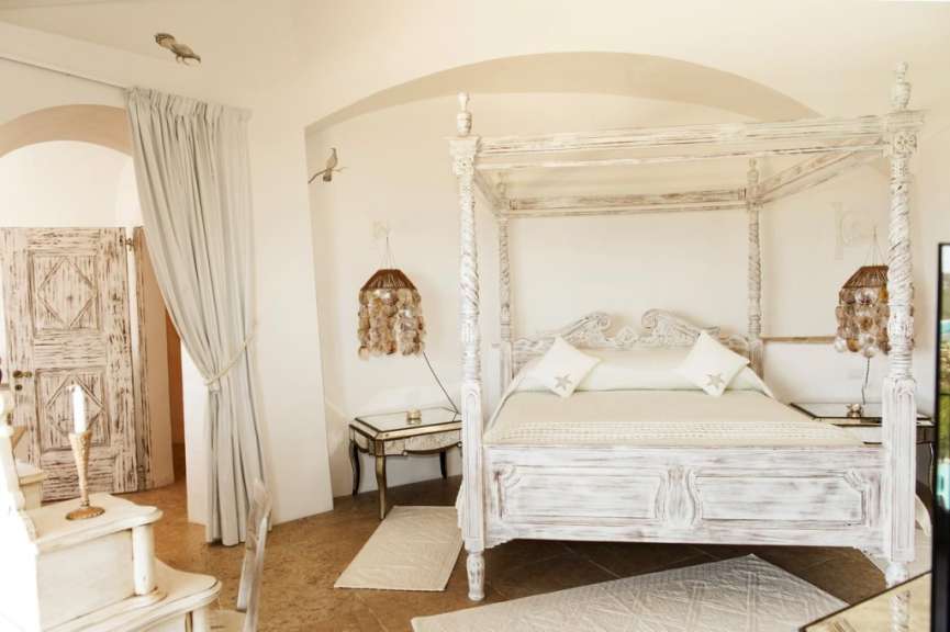 Ein romantisches Himmelbett mit verschnörkelten Säulen steht in einem hellen Schlafzimmer mit weißen Wänden und einem Steinboden.