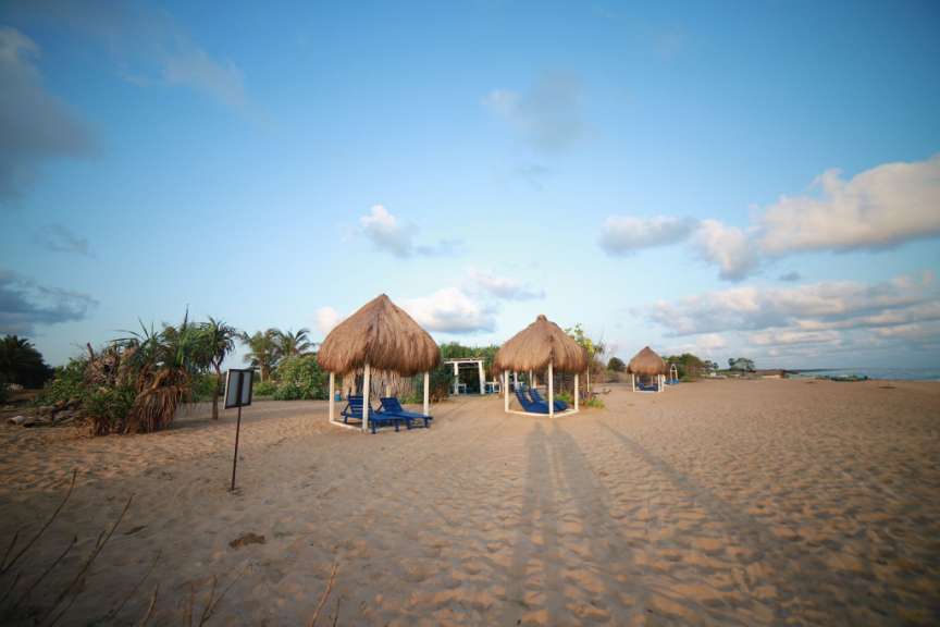 Ein Strand mit Strohhütten, Liegestühlen und Palmen unter einem blauen Himmel mit Wolken.