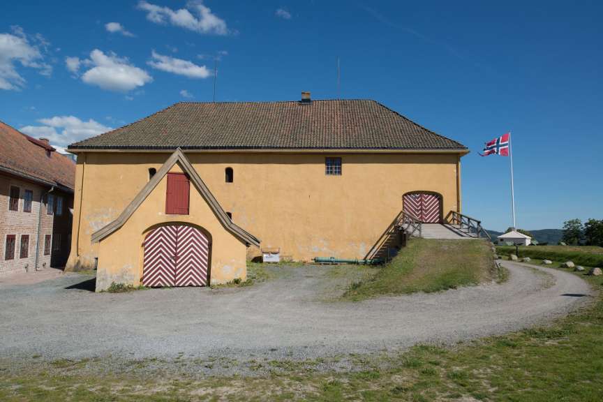 Ein gelbes Gebäude mit roten Türen steht auf einem Hügel vor blauem Himmel mit Wolken, daneben weht eine norwegische Flagge.