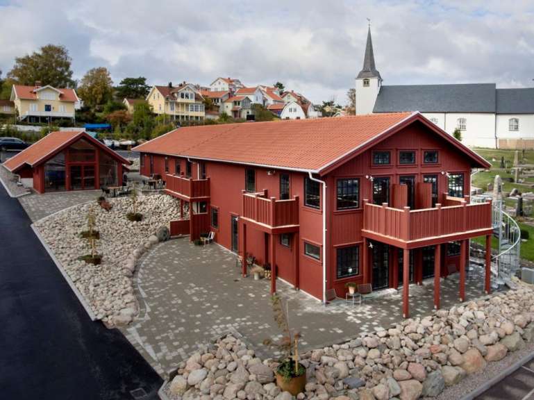 Ein zweistöckiges rotes Holzhaus mit Balkonen und großen Fenstern steht auf einem felsigen Grundstück mit einem gepflasterten Hof.
