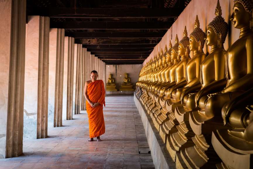Ein Mönch steht in einem Flur mit vielen goldenen Buddhastatuen