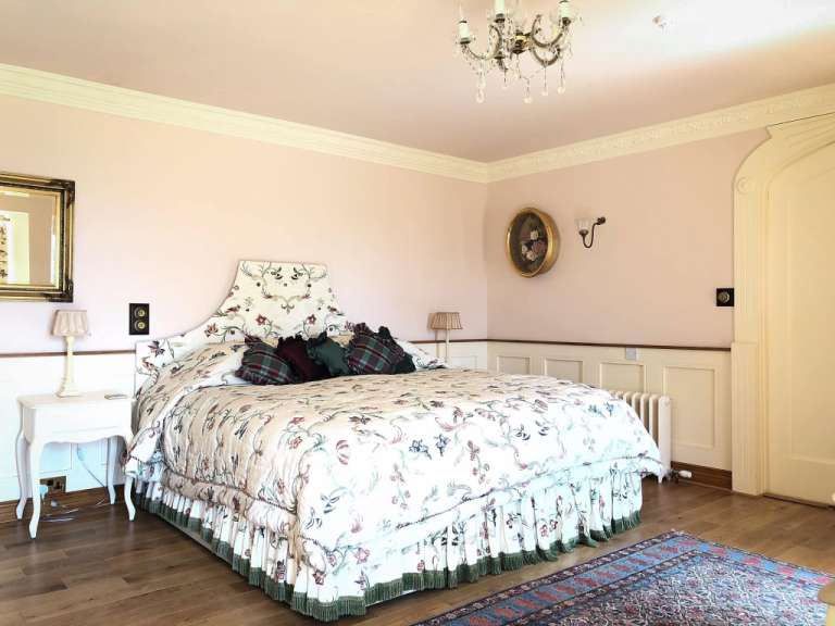 Ein reich verziertes Bett mit floralem Muster, bedeckt mit einer Tagesdecke und Kissen, steht in einem pastellfarbenen Schlafzimmer mit Holzboden und einem orientalischen Teppich.