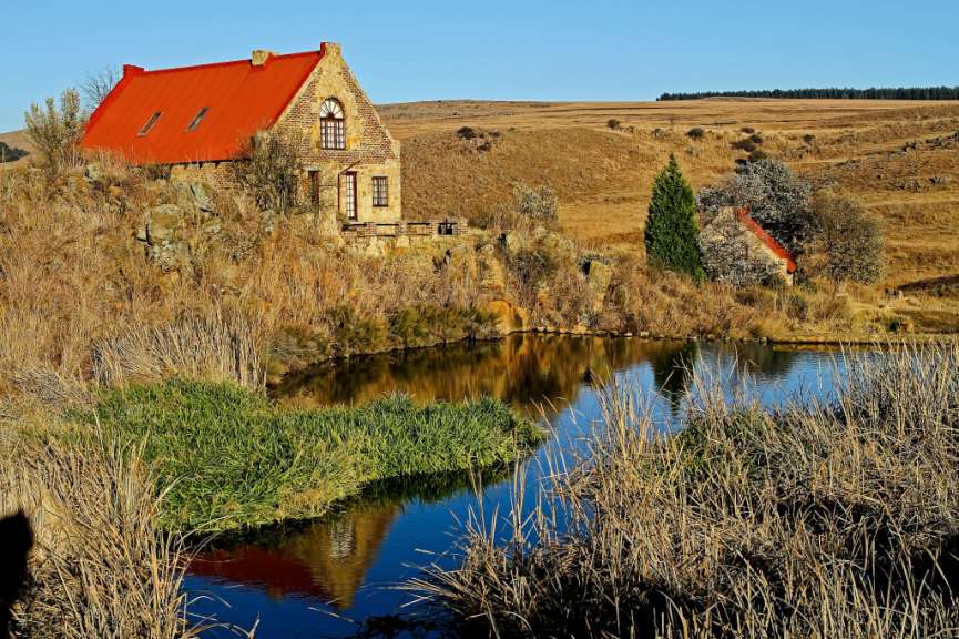 Ein Steinhaus mit rotem Dach steht an einem Teich, umgeben von trockenem Gras und grünen Pflanzen.