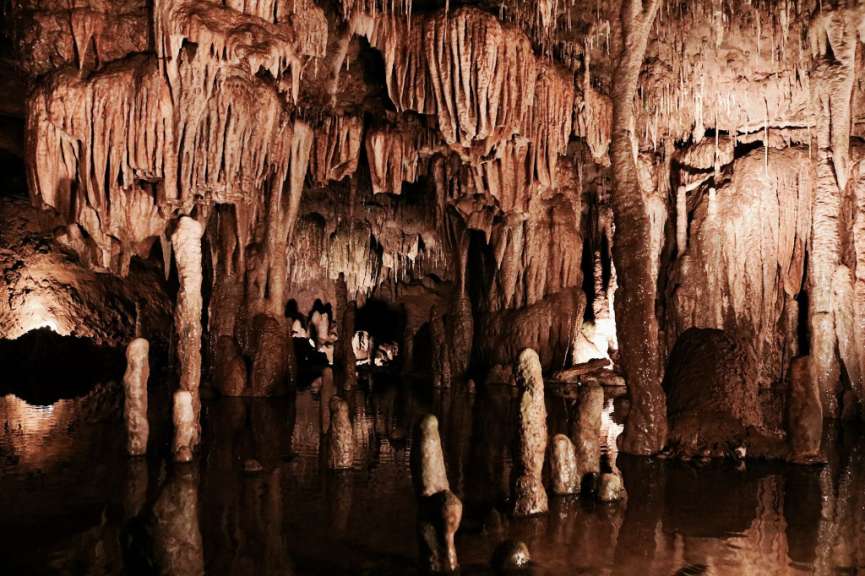 Diese Aufnahme zeigt eine Höhle mit vielen Stalaktiten und Stalagmiten, die sich in dunklem Wasser spiegeln.