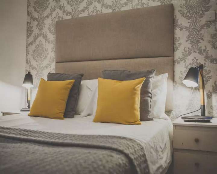 Ein Bett mit einer großen Kopfstütze, zwei gelben und zwei grauen Kissen sowie zwei Nachttischlampen steht vor einer gemusterten Tapete.