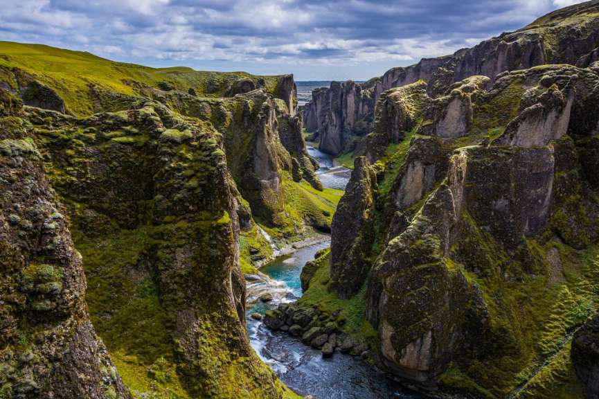 Ein Fluss fließt durch eine üppig grüne, moosbewachsene Schlucht unter einem bewölkten Himmel.