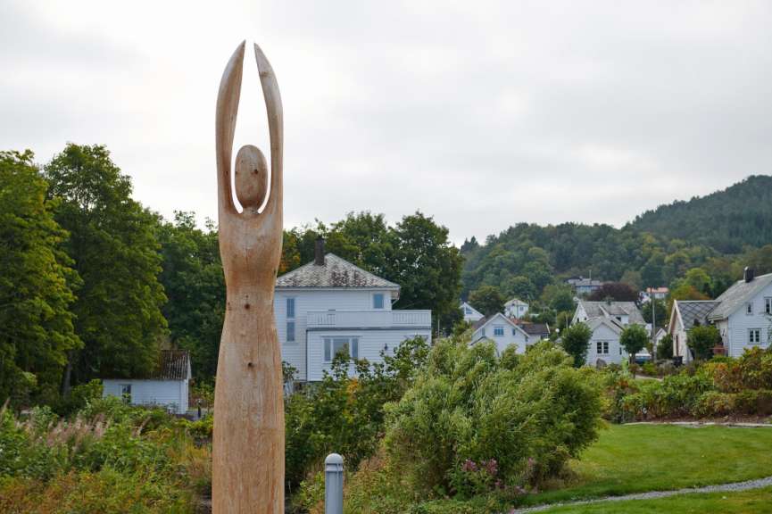 Ein holzgeschnitztes Kunstwerk, das eine Figur mit erhobenen Armen darstellt, steht im Vordergrund vor einer Reihe von weißen Häusern und einem bewaldeten Hügel.