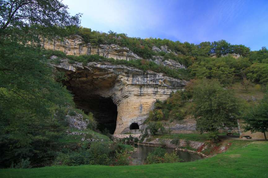 Ein großer Höhleneingang erstreckt sich über eine Felswand, umgeben von Bäumen und Vegetation, mit einem Fluss im Vordergrund.