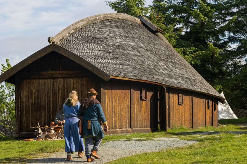 Zwei Personen in historischer Kleidung gehen auf ein Holzgebäude mit einem gekrümmten Dach zu.